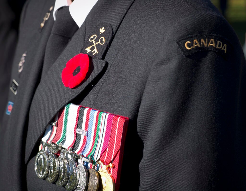 A red poppy pinned to the lapel of a dark Canadian Forces uniform, above a row of service medals and beside a Canada shoulder flash.