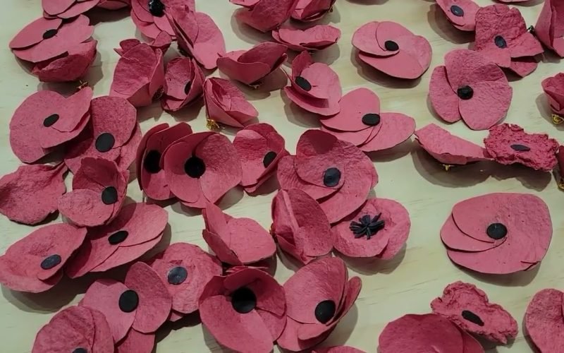 Dozens of red seed paper poppies with black centres laid out on a pale wooden table, some assembled and some still flat.