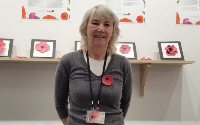 Wendy Sperry, founder of The Poppy Project, wearing a seed paper poppy and standing in front of a wall display of framed poppies.